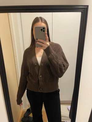 Brun ribbstickad kofta med knappar - Säljer en mysig brun kofta med ribbstickat material och stora knappar framtill. Koftan har v-ringning, långa ärmar och en avslappnad passform. 