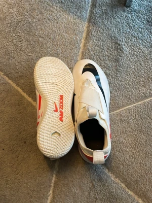Nike futsalskor - Säljer ett par Nike Zoom futsalskor i vitt med röda och svarta detaljer. Skorna har en slimmad design med kardborreband över vristen och slitstark gummisula för bra grepp på planen. Perfekta för dig som vill ha stilrena och funktionella skor till inomhusfotboll. Dom är i bra skick och är i storlek 38,5.