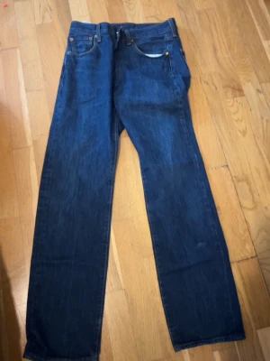 Levi's raka blå jeans w30 - Säljer ett par klassiska blå jeans från Levi's med rak passform. Jeansen har fem fickor, synliga nitar och Levi's ikoniska läderpatch bak i midjan. Tillverkade i slitstark denim med mörkblå tvätt och diskreta sömmar. Perfekta för en avslappnad och tidlös stil.