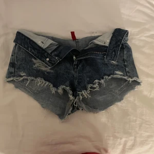 Blå lågmidjade shorts med fransar - Säljer ett par snygga blå jeansshorts med fransiga kanter och slitna detaljer. Shortsen har en dragkedja och knapp framtill samt fickor både fram och bak. Perfekta för en avslappnad sommarlook!