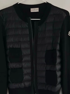 Svart dunväst från Moncler - Snygg svart dunväst från Moncler med quiltad framsida, två framfickor och dragkedja. Västen har rund halsringning och Moncler-logga på ärmen. Materialet är en mix av dun och textil, vilket ger en modern och sportig look.