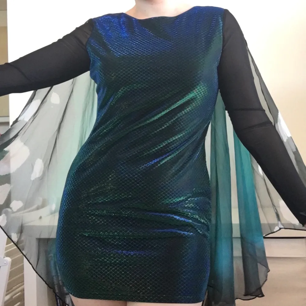 The label is cut off but it fits size M/L. The length from neck down is size 75cm. Dress has been used twice, and one of the arms has been a little damaged around the hand/wing, but it seems easy enough to sew together! The dress has a black colour with shimmering dark blue and dark green. The arms are half transparent and the wrists connect decorated butterly 