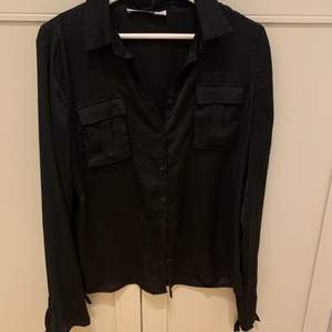 En svart blus/skjorta i satin från NAKD. Den har runt handlederna band som sitter fast för att knyta rosett. Den är i storlek 36. 