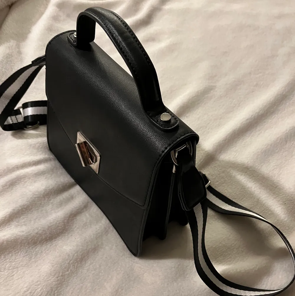 Snygg, svart, stilren handväska med ett ett randigt band som piffar upp den och outfiten! . Accessoarer.