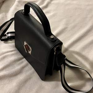 Snygg, svart, stilren handväska med ett ett randigt band som piffar upp den och outfiten! 
