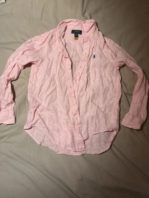 Ljusrosa linneskjorta Polo Ralph Lauren 10-12 år - Säljer en ljusrosa skjorta från Polo Ralph Lauren i luftigt linne. Skjortan har klassisk krage, knappar framtill och den ikoniska blå loggan broderad på bröstet. Perfekt för en avslappnad men ändå stilren look.