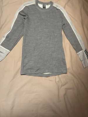 Grå underställströja från Helly Hansen - Säljer en grå långärmad tröja från Helly Hansen med vita detaljer längs ärmarna och diskreta svarta ränder på ena ärmslutet. Tröjan är tillverkad i ett funktionsmaterial som andas, perfekt för träning eller utomhusaktiviteter. Finns annons på matchande byxor.