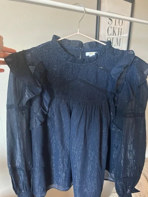 Mörkblå volangblus Gina Tricot - Mörkblå blus från Gina Tricot med långa ärmar, volangdetaljer och smock upptill. Materialet är lätt transparent på ärmarna, med subtilt glittrigt skimmer och har en hög krage med rynk. Perfekt för festliga tillfällen som jul och nyår! Använd någon enstaka gång under nyår.