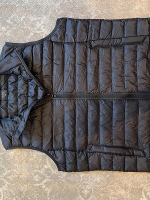Svart dunväst från Everest - Snygg svart dunväst från Everest med quiltad design och dragkedja framtill. Västen har två sidofickor och hög krage för extra värme. Perfekt att slänga över en hoodie eller tröja när det är kyligt ute.