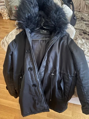 Svart parkas med fuskpäls från K-Zell - Snygg svart parkas från K-Zell med stor huva och fuskpäls. Jackan har dragkedja och knappar framtill, samt snörning i midjan för bättre passform. Perfekt för kalla dagar och riktigt mysig att bära.