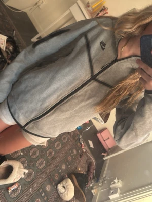 Grå zip hoodie från Nike - Finns matchade byxor med!! Säljer en grå zip hoodie från Nike med svarta detaljer och dragkedja framtill. Hoodien har en huva och Nike-logga på bröstet. Perfekt för en avslappnad och sportig stil. Materialet känns mjukt och bekvämt.
