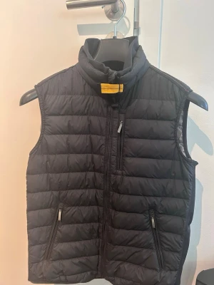 Svart dunväst med hög krage - Snygg svart dunväst från Parajumpers med hög krage och gul detalj vid halsen. Västen har tre dragkedjeförsedda fickor framtill och quiltad design. 