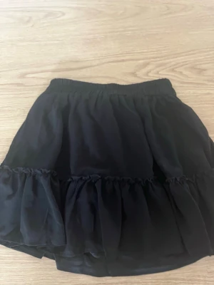 Svart volangkjol med resårmidja - Säljer en svart kortkjol med volangdetalj och resår i midjan. Kjolen har en luftig och flowy känsla, perfekt för dig som gillar en enkel men ändå söt stil. Materialet känns lätt och passar bra till olika outfits.