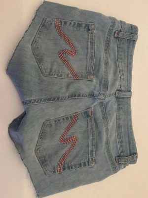 Ljusblå jeansshorts med broderi - Säljer ett par ljusblå jeansshorts med rå kant och röda broderade detaljer på bakfickorna. Shortsen har bälteshällor, dubbla knappar framtill och klassisk femficksdesign. Perfekta för sommaren och ger en avslappnad vibe.