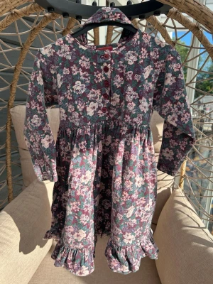 Klänning strl. 98/104 - Långärmad klänning från Tuffer med blommigt mönster i lila, rosa och grönt. Klänningen har knappar framtill, volangkant nedtill och mjukt tyg som känns bekvämt.