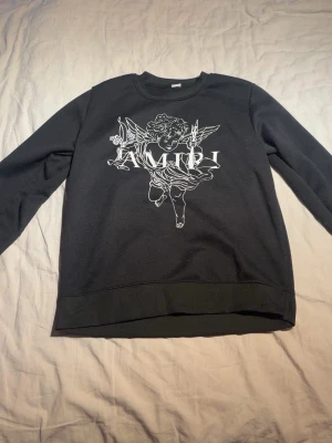 Svart Amiri sweatshirt - Svart sweatshirt från AMIRI med ett stort vitt tryck av en ängel och texten 'AMIRI' på bröstet. Tröjan har rund halsringning, långa ärmar och ribbade muddar. Perfekt för dig som gillar streetwear och snygga prints.