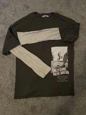 Mörkgrö långärmad topp från H&M med tryck - Cool svart långärmad topp från H&M med kontrasterande vita ärmar och tryck framtill med texten 'ONLY COOL DAYS AHEAD' och en bild på en cyklist. Oversized passform och skön bomullskänsla. Perfekt för dig som gillar streetwear vibes.