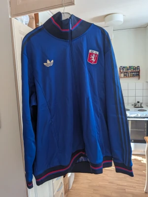Blå Adidas track jacket OL - Snygg blå track jacket från Adidas med OL-logga på bröstet och klassiska svarta ränder längs ärmarna. Dragkedja framtill, ribbade muddar och krage med röd och blå detalj. Perfekt för en sportig och retro look.