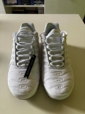 Vita Nike Air Max Plus sneakers - Säljer ett par helvita Nike Air Max Plus sneakers med ikoniska vågmönster och synlig Air-dämpning i sulan. Skorna har mesh- och syntetmaterial på ovandelen, samt vita snören och detaljer. Perfekta för dig som gillar en clean och sportig look.
