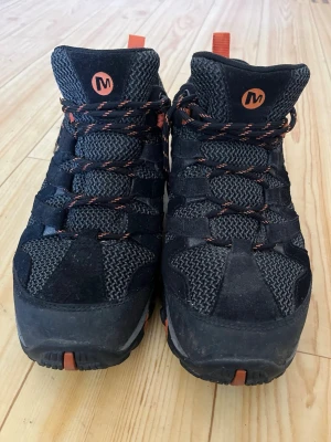 Svarta Merrell kängor med orange detaljer - Säljer ett par svarta kängor från Merrell med orange detaljer och snörning. Skorna har robust sula, meshpaneler för ventilation och förstärkta partier i mocka. Perfekta för utomhusaktiviteter och äventyr. Snygg och sportig design med Merrell-logga på sidan.