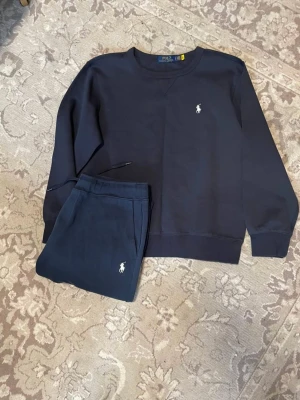 Marin blå track suit från Polo - Mörkblått mjukis set från Polo Ralph Lauren med matchande sweatshirt och byxor. Båda plaggen har den klassiska vita Polo-loggan broderad på bröstet och benet. Mjukt material och avslappnad passform, perfekt för chill dagar hemma.