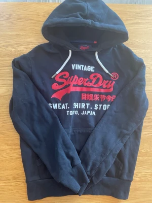 Mörkblå Superdry hoodie med tryck - Snygg mörkblå hoodie från Superdry med stort rött och vitt tryck på bröstet och japanska tecken. Klassisk huva med vita snören och känguruficka framtill. Perfekt för en avslappnad streetwear-look.