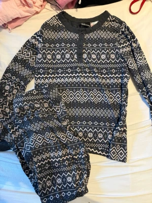 Mörkgrå pyjamas med mönster - Mysig tvådelad pyjamas i mörkgrått med vitt geometriskt mönster. Tröjan har rund halsringning, knappar framtill och långa ärmar. Byxorna har samma mönster och ser ut att vara i mjukt bomullsmaterial. Perfekt för chillkvällar hemma.