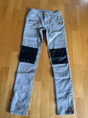 Grå jeans från Kappahl - Snygga grå jeans med svarta paneler över knäna och dragkedjor vid bensluten. Fickor med dekorativa nitar och stjärnor baktill samt coola sömmar. Perfekta för dig som vill sticka ut lite extra.