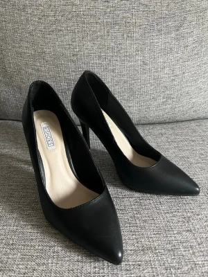 Svarta pumps från Nelly - Stilrena svarta pumps från Bianco med spetsig tå och hög klack. Skorna har en slät yta i konstläder och en ljus innersula. Perfekta för dig som gillar en tidlös och elegant look. Använda 1 gång inomhus.