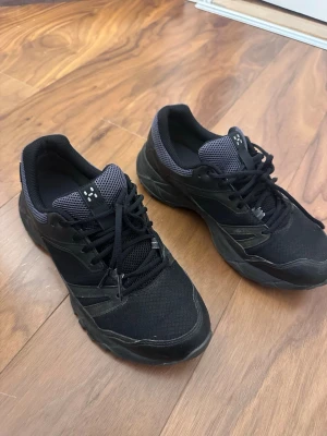 Svarta sneakers med mesh och detaljer - Snygga svarta sneakers med meshpartier och svarta syntetdetaljer. Skorna har svart snörning och en tjock sula för extra komfort. Insidan är fodrad med grått tyg och tungan har ett diskret mönster. Perfekta för dig som gillar en sportig och stilren look.