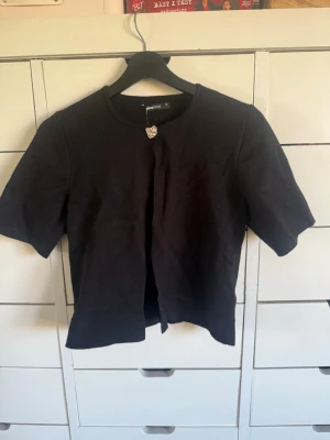 Svart kortärmad topp med knapp - En svart, kortärmad topp med rund hals och en knapp framtill. Toppen har en enkel och stilren design och är tillverkad i ett stickat material som ger en mjuk känsla. Perfekt för dig som gillar minimalistisk stil.