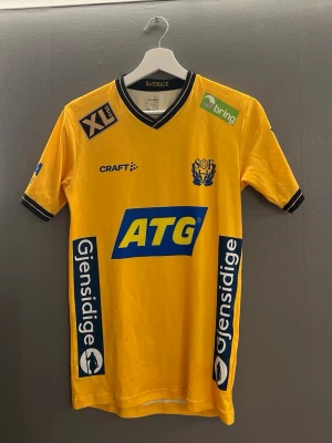  Sveriges handbollslandslagströja  - Gul matchtröja med svarta detaljer från Craft, prydd med flera sponsorer som ATG, Gjensidige och Sparbanken Skåne. Tröjan har V-ringning, korta ärmar och klubbmärke på bröstet. Perfekt för dig som gillar sportig stil och vill sticka ut på planen.