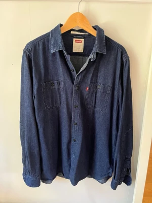 Mörkblå jeansskjorta från Levi's - Snygg mörkblå jeansskjorta från Levi's med klassisk krage, två bröstfickor och knappar framtill. Skjortan har långa ärmar och en relaxed passform. Perfekt att bära öppen eller stängd för en avslappnad look.