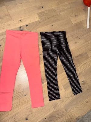 Två leggings från Lupilu, strl 98/104 - Säljer två par leggings från Lupilu i storlek 98/104. Ett par är helrosa och det andra är svart med tunna vita och rosa ränder. Båda har elastisk midja och är gjorda i mjukt bomullsmaterial, perfekta för en bekväm och avslappnad stil. Två leggings från Lupilu, strl 98/104