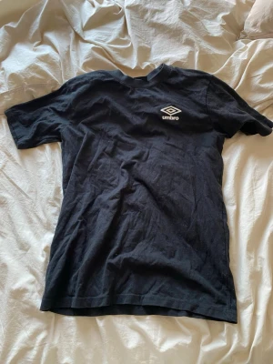 Svart t-shirt från Umbro - Säljer en svart t-shirt från Umbro med klassisk logga tryckt på bröstet. T-shirten har rund hals och korta ärmar, tillverkad i mjuk bomull som känns skön mot huden. Perfekt för en sportig och avslappnad stil.