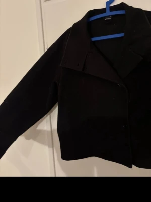 Kort svart kappa med bred krage - Stilren kort svart kappa med bred krage och knäppning framtill. Kappan har långa ärmar och en klassisk, rak passform. Perfekt för dig som gillar minimalistisk och tidlös stil.köpt för 600kr, säljer för 300kr. Pris kan diskuteras.