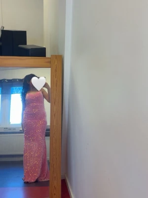 Rosa glittrig långklänning - Säljer en superfin rosa långklänning med massor av glitter och smala axelband. Klänningen har en figurnära passform och är perfekt för dig som vill sticka ut. Materialet känns mjukt och skimrande, och den har en klassisk siluett som ger en elegant look.Klänningen är i väldigt fint skick! Det finns endast lite lätt mörkare slitage längst ner på fållen/kanten från när man gått med den, vilket är helt oundvikligt på maxiklänningar. Det är ingenting som syns alls när man har på sig den! 