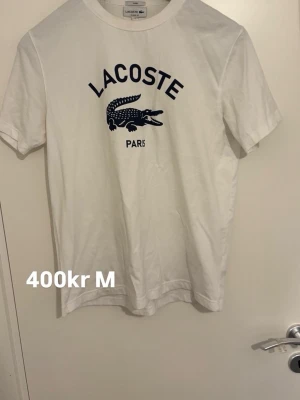 Vit Lacoste t-shirt med tryck - Vit t-shirt från Lacoste med klassiskt blått krokodiltryck och texten 'Lacoste Paris' på bröstet. T-shirten har rund halsringning och korta ärmar. Tillverkad i mjuk bomull för en skön känsla.