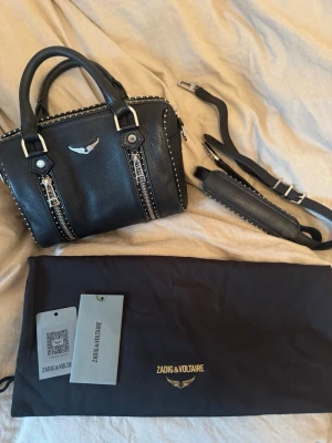 Svart handväska Zadig & Voltaire sunny bag - Snygg svart handväska i läder från Zadig & Voltaire med silverfärgade detaljer, dubbla dragkedjor framtill och dekorativa nitar längs kanterna. Väskan har både handtag och avtagbar axelrem samt märkets ikoniska vinglogga i metall på framsidan. Dustbag och tags med serienummer medföljer. Väldigt sparsamt använd och inprincip inga tecken på användning. Köpt från Zalando förra året för 5000 kr. Kan gå ner lite i pris vid snabb affär!💗Kontakta mig gärna om ni har ytterligare frågor💕