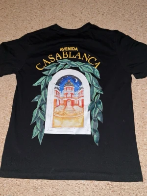 Svart t-shirt med Casablanca-tryck - Svart t-shirt med färgglatt tryck av en byggnad och gröna blad, samt texten 'Avenida Casablanca' på ryggen och mindre tryck framtill. Klassisk passform och rund hals. Perfekt för dig som gillar unika motiv och streetstyle.