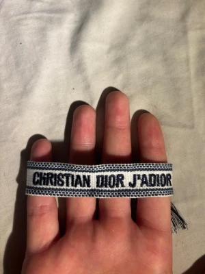Christian Dior armband - Snyggt vävt armband från Christian Dior med texten 'J'Adior' i svart på vit botten. Armbandet har en bred design och avslutas med dekorativa tofsar. Perfekt accessoar för att lyfta din outfit med en exklusiv touch.