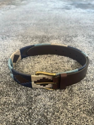 Brunt och blått mönstrat bälte - Snyggt bälte i brunt skinn med detaljer i blått, beige och mörkblått. Bältet har ett unikt geometriskt mönster och ett klassiskt metallspänne. Perfekt för att lyfta din outfit med lite extra färg och stil.