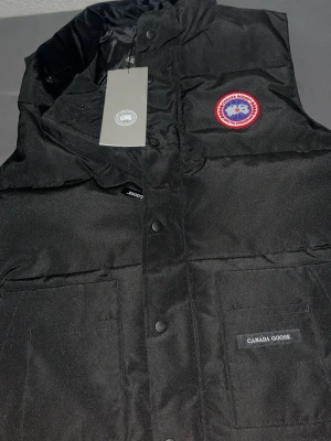 Svart dunväst från Canada Goose - Snygg svart dunväst från Canada Goose med klassisk logotyp på bröstet och två stora fickor framtill. Västen har hög krage och tryckknappar. Perfekt för lager-på-lager och håller dig varm under kyliga dagar.