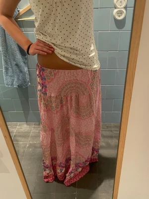 Långkjol i rosa mönster från MARTA - Säljer en flowy långkjol från MARTA i ett färgglatt rosa mönster med inslag av blått, orange och vitt. Kjolen har smock i midjan och ett bohemiskt, paisley-inspirerat tryck. Perfekt för dig som gillar en avslappnad och färgstark stil.