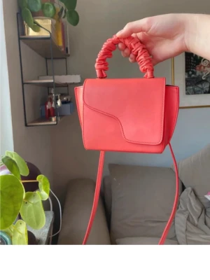  Korall väska  från ATP Atelier - Trendig korall handväska från ATP Atelier i slätt läder. Väskan har en unik form med asymmetrisk lock och ett scrunchie-inspirerat handtag samt lång axelrem. Perfekt för dig som vill sticka ut med en färgklick och cool design. 