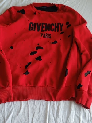 Röd distressed Givenchy hoodie - Säljer en röd hoodie från Givenchy med svart text 'GIVENCHY PARIS' på bröstet. Hoodien har ett distressed, slitet utseende med flera hål och slitningar över hela plagget. Tillverkad i mjukt bomullsmaterial och har ribbade muddar vid ärmslut och nederkant.