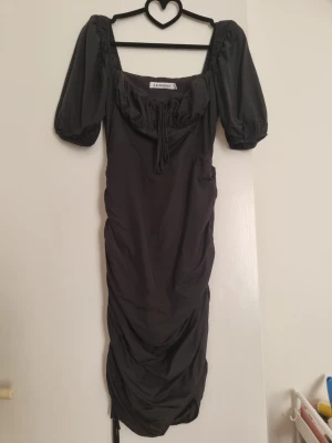 Svart klänning med puffärm från Adoore - Trendig svart klänning i modellen Palermo från Adoore med puffärmar och snörning vid bysten. Klänningen har en åtsittande passform, rynkade detaljer längs kjolen och fyrkantig urringning. Perfekt för dig som vill ha en stilren och modern look.