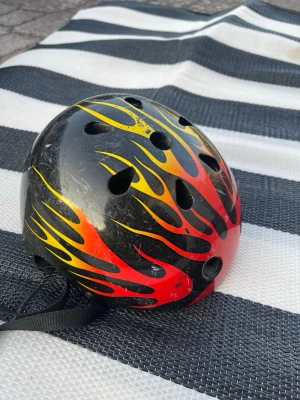 Cykelhjälm med eldflammor - Säljer en cool cykelhjälm i svart med röda och gula eldflammor. Hjälmen har flera ventilationshål och justerbart hakband. Perfekt för dig som vill sticka ut på cykeln eller skateboarden.