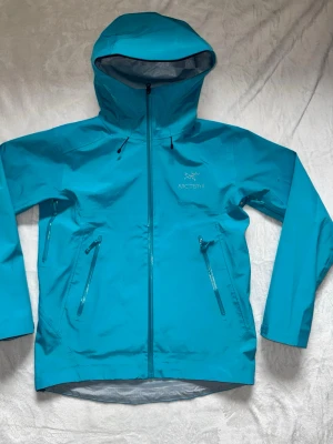 Blå vindjacka från Arc'teryx - Snygg blå vindjacka från Arc'teryx med huva och hel dragkedja framtill. Jackan har två snedställda fickor med dragkedja och justerbar huva. Tillverkad i ett lätt och slitstarkt material som står emot vind och regn. Perfekt för utomhusaktiviteter. Dragkedjan är lite seg men funkar ändå.