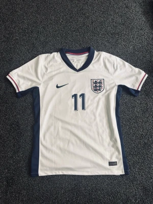 England matchtröja Nike nr 11 Foden - Säljer en vit England fotbollströja från Nike med marinblå detaljer, nummer 11 och namnet Foden på ryggen. Tröjan har korta ärmar, broderad landslagssköld och V-ringad krage med röd och blå kant. Perfekt för dig som älskar fotboll och landslaget.